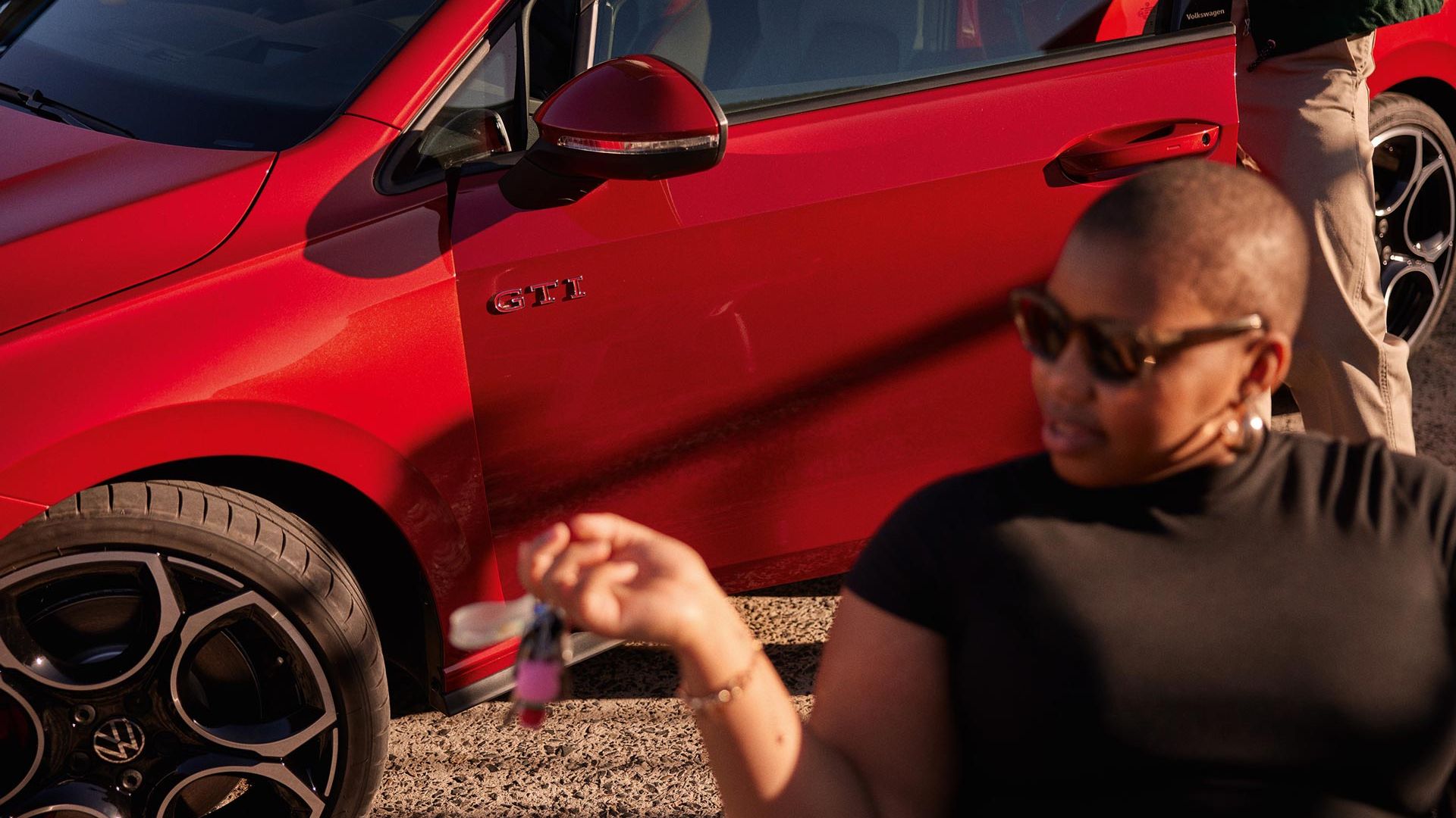 Detailní pohled na dveře řidiče vozu Volkswagen Golf GTI v červené barvě. Žena odchází od vozu.