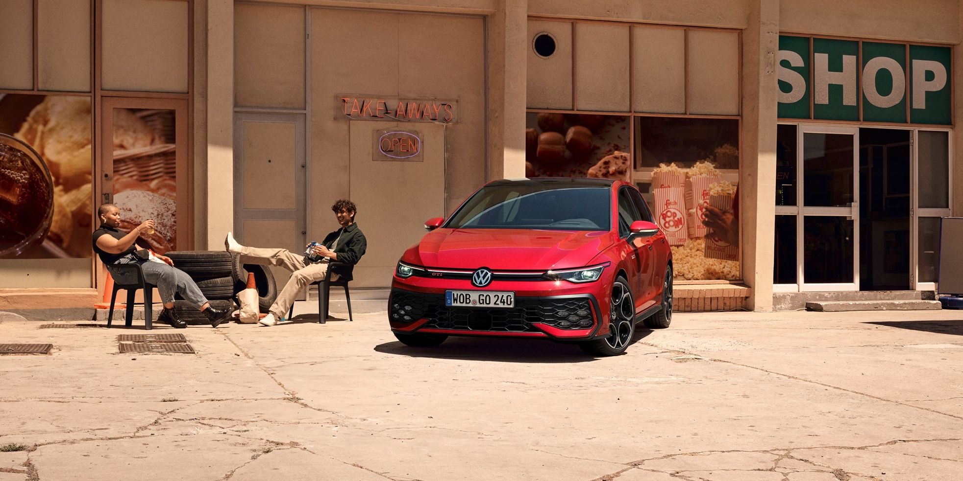 Volkswagen Golf GTI v červené barvě stojí před budovou. Vedle vozu sedí dvě osoby.