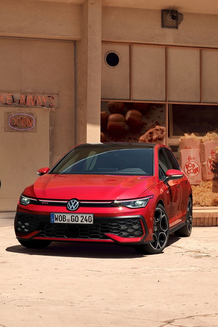 Volkswagen Golf GTI v červené barvě stojí před budovou. Vedle vozu sedí dvě osoby.