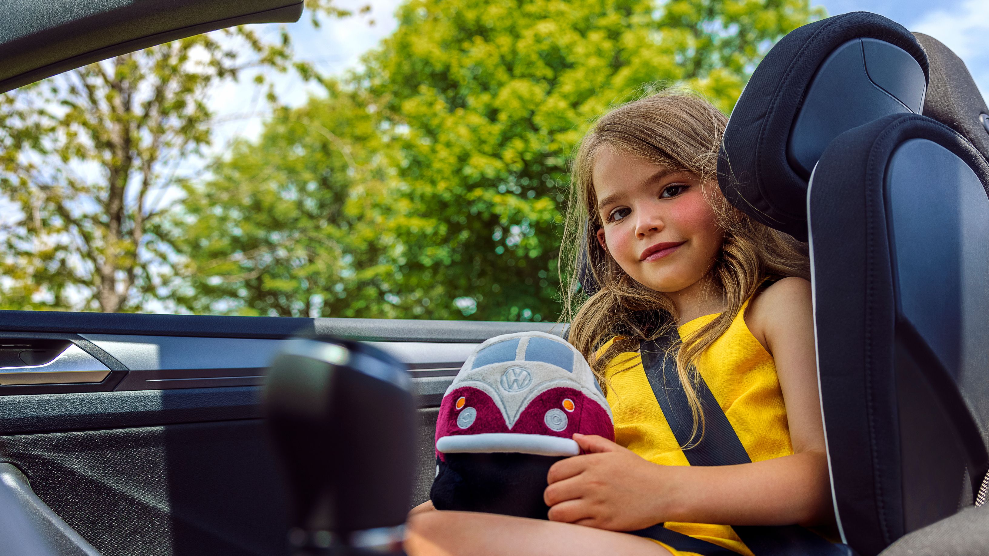 Ein Mädchen sitzt in einem VW Kindersitz und hat ein VW Bulli Kuscheltier iin der Hand